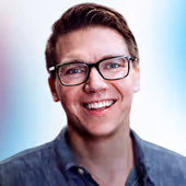 Smiling man with glasses and short hair wearing a blue button-up shirt against a blurred background.