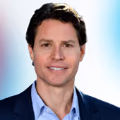 Man with dark hair wearing a blue suit and light blue shirt against a blurred background.