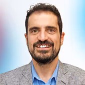 Smiling man with dark curly hair, beard, and mustache, wearing a gray blazer and blue shirt.
