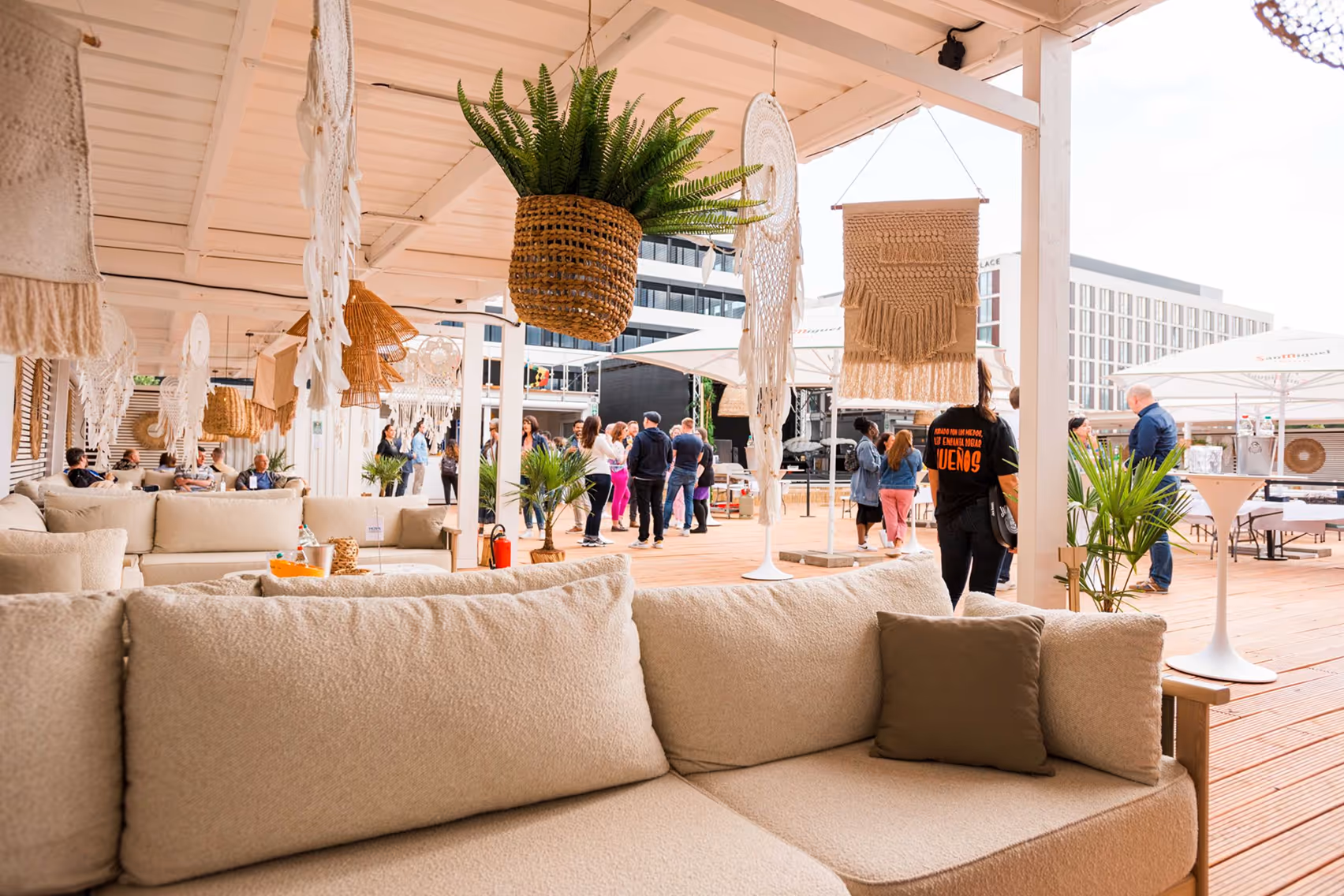 Helle Lounge mit beigen Sofas, hängenden Pflanzenkörben und Makramee-Dekoration, Menschen im Hintergrund bei einem Event im Freien.