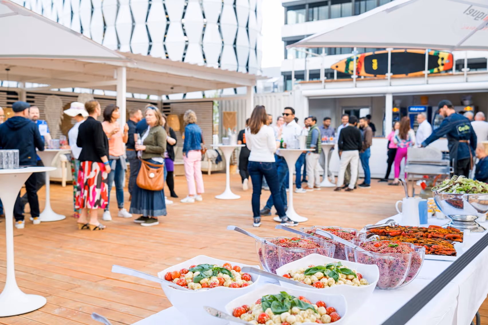 Outdoor-Event mit Menschen, die an Stehtischen stehen, davor ein Buffettisch mit Salaten und Vorspeisen.