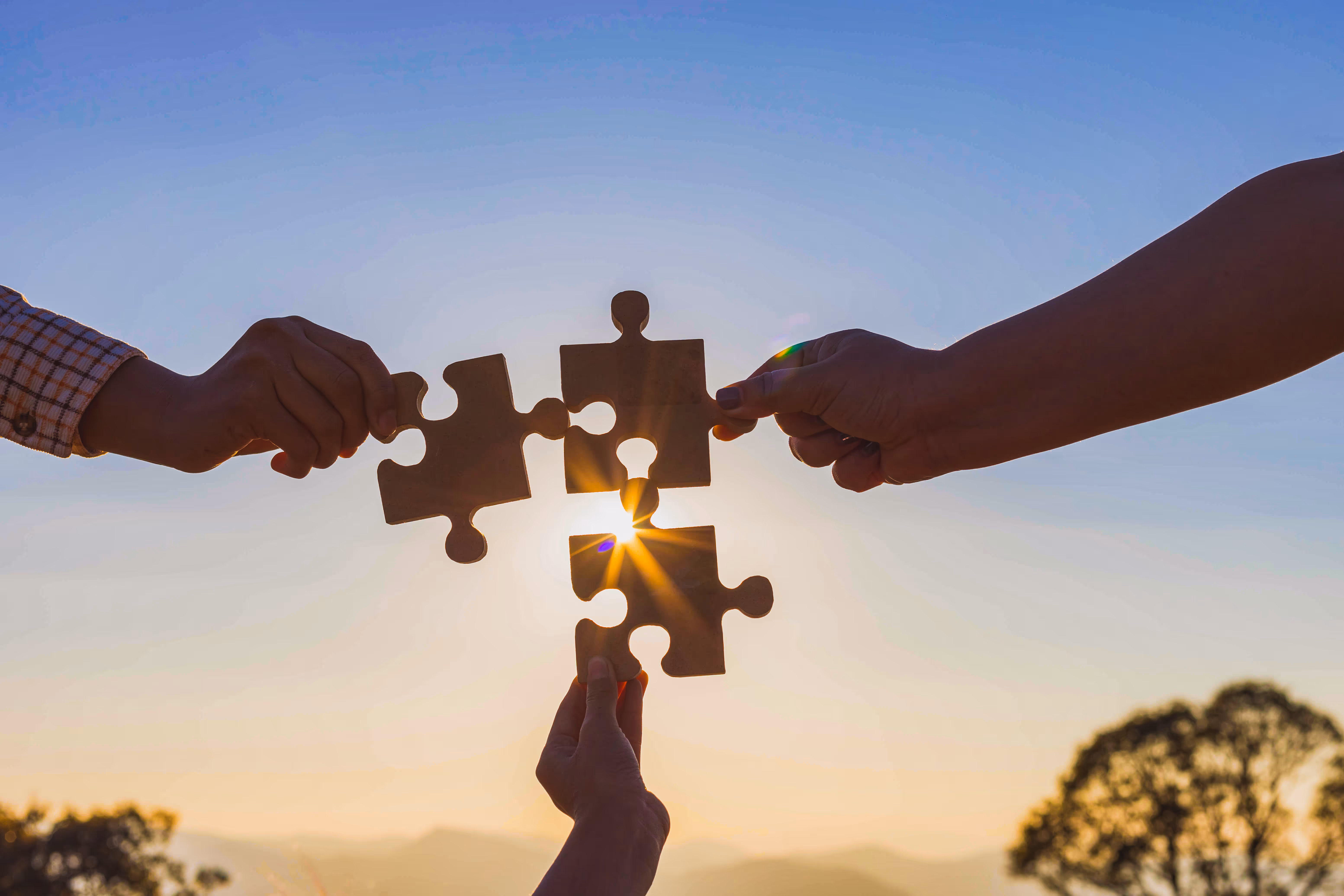 Three hands holding and connecting four wooden puzzle pieces with the sun shining through them at sunset.