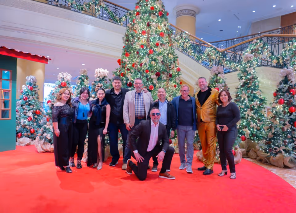 Group of ten adults posing on a red carpet in front of decorated Christmas trees and festive ornaments inside a building.