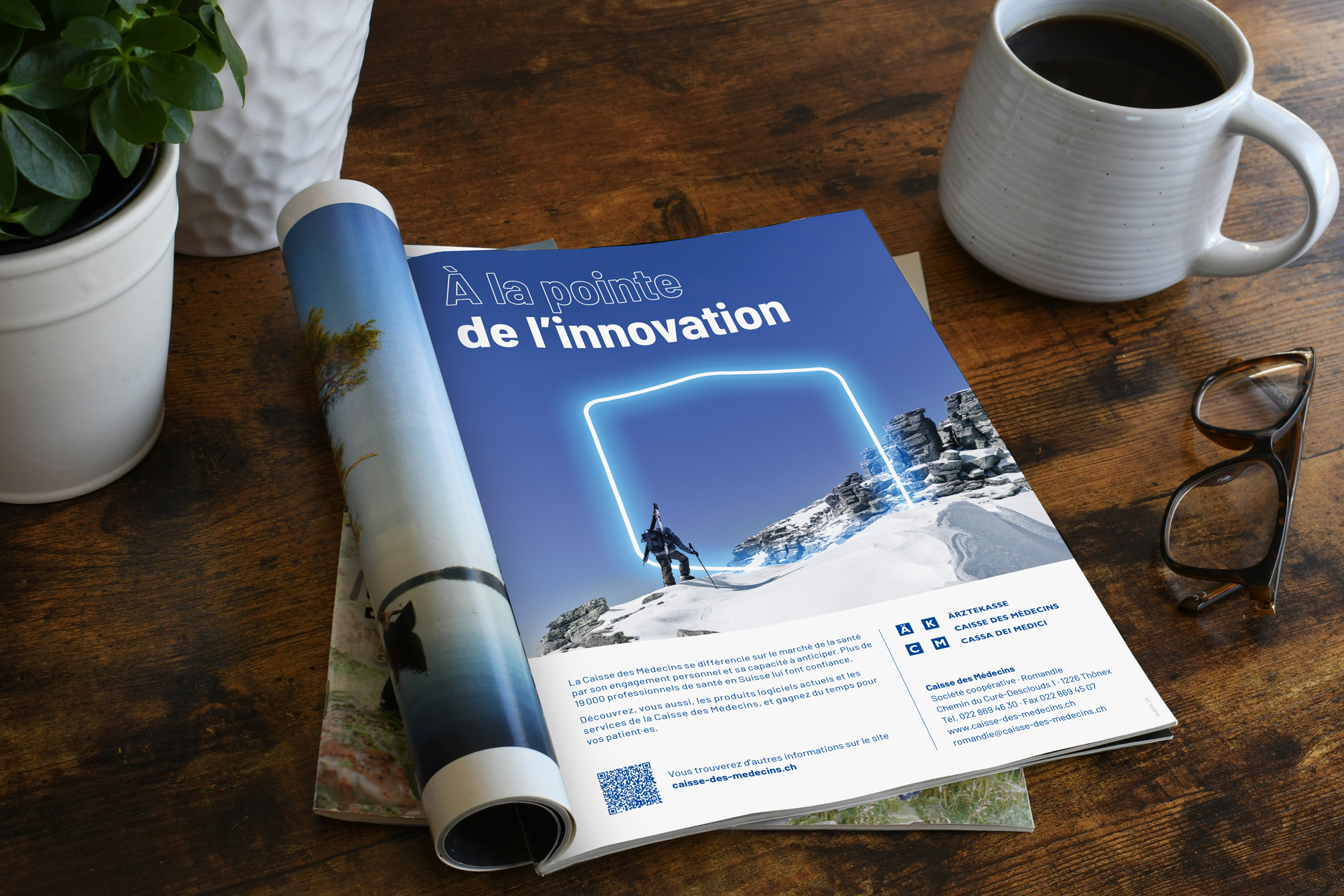 Aufgeschlagene Zeitschrift mit französischer Ärztekasse Genossenschaft Anzeige, Schneelandschaft und Skifahrer unter einem leuchtenden blauen Rahmen, daneben Kaffeetasse, Brille und Pflanze auf Holztisch.