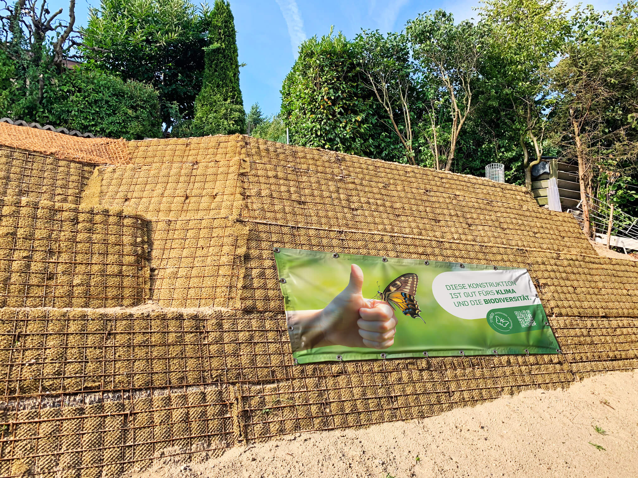 Hangschutzkonstruktion aus Stützgitter mit Naturvlies, davor ein Banner mit einem Daumen hoch, einem Schmetterling und dem Text: Diese Konstruktion ist gut fürs Klima und die Biodiversität.