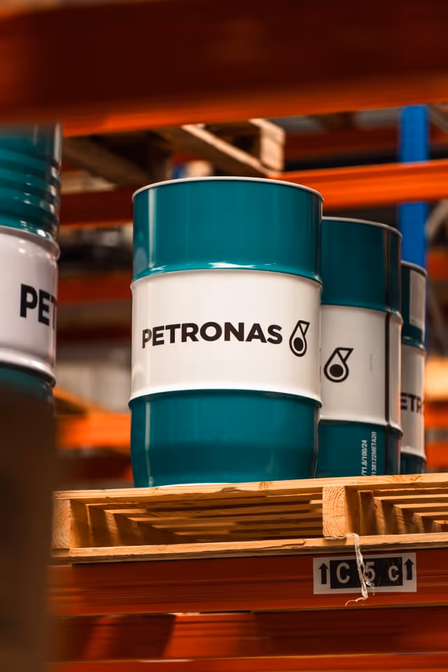 Metalen vaten in groen en wit met het Petronas-logo, op houten pallets in een magazijn.