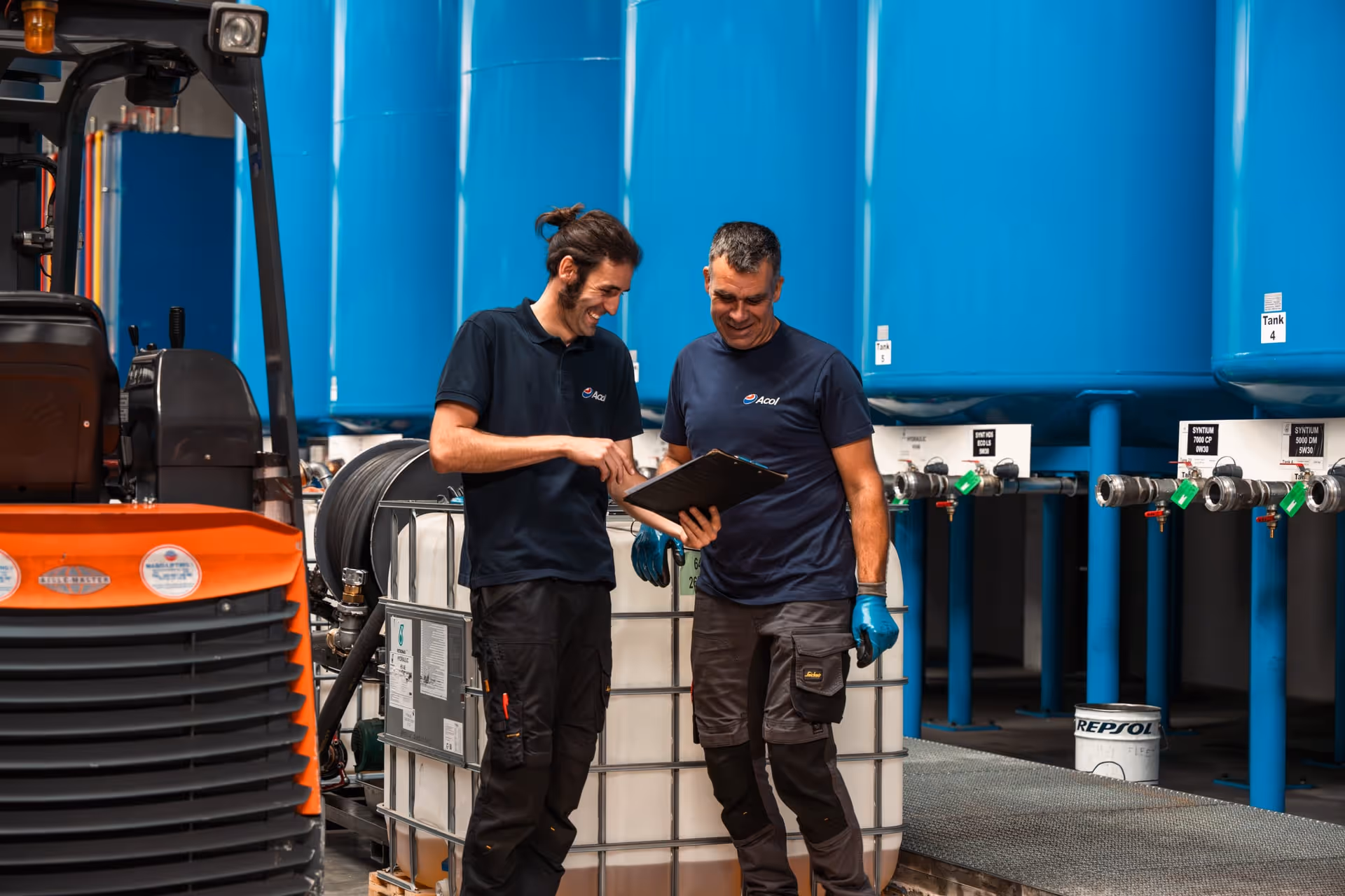 Twee werknemers in marineblauwe overhemden staan bij grote blauwe tanks en een oranje vorkheftruck te praten over een klembord.