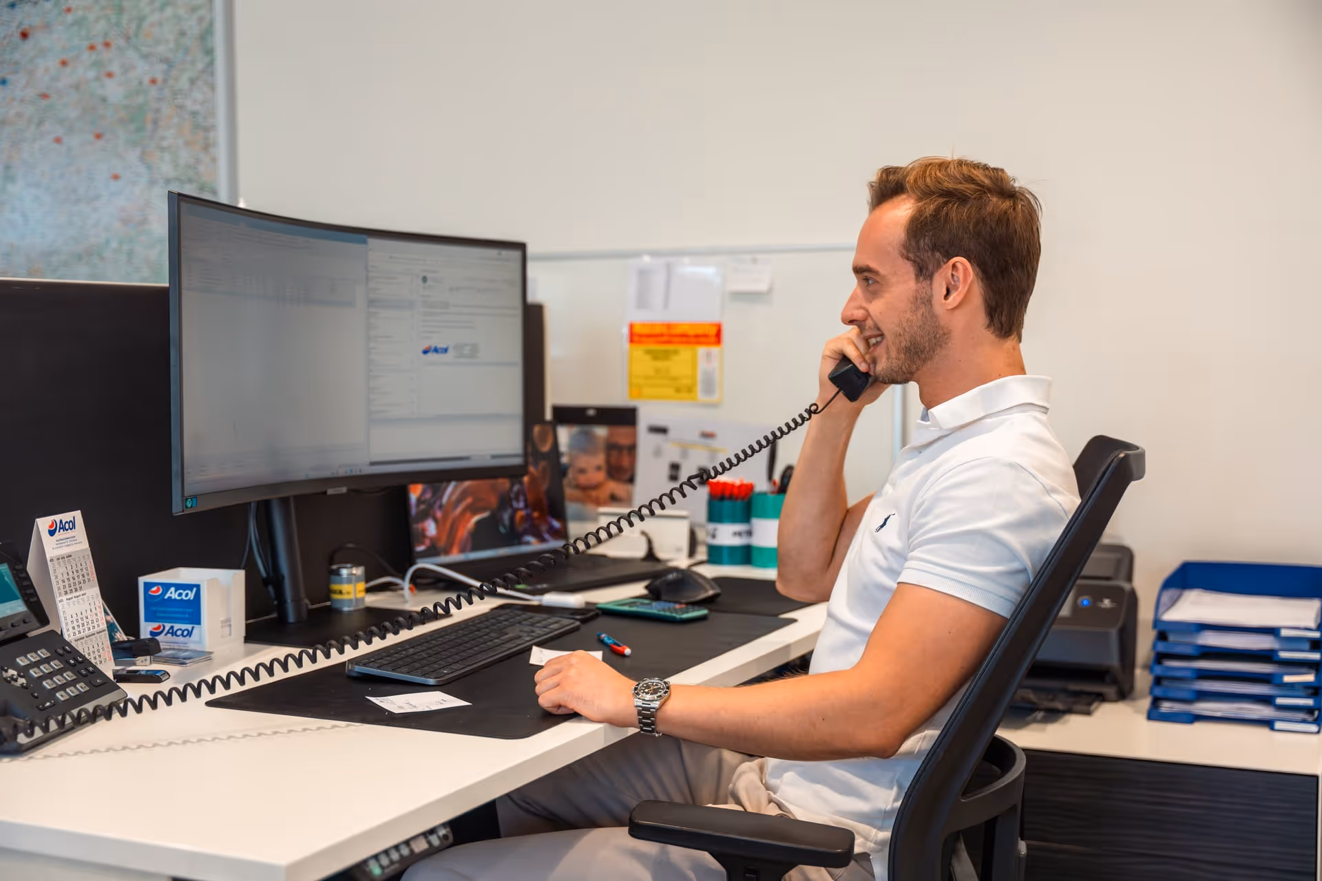 Een man in een wit poloshirt die aan een bureau met computermonitors en kantoorbenodigdheden zit te bellen.