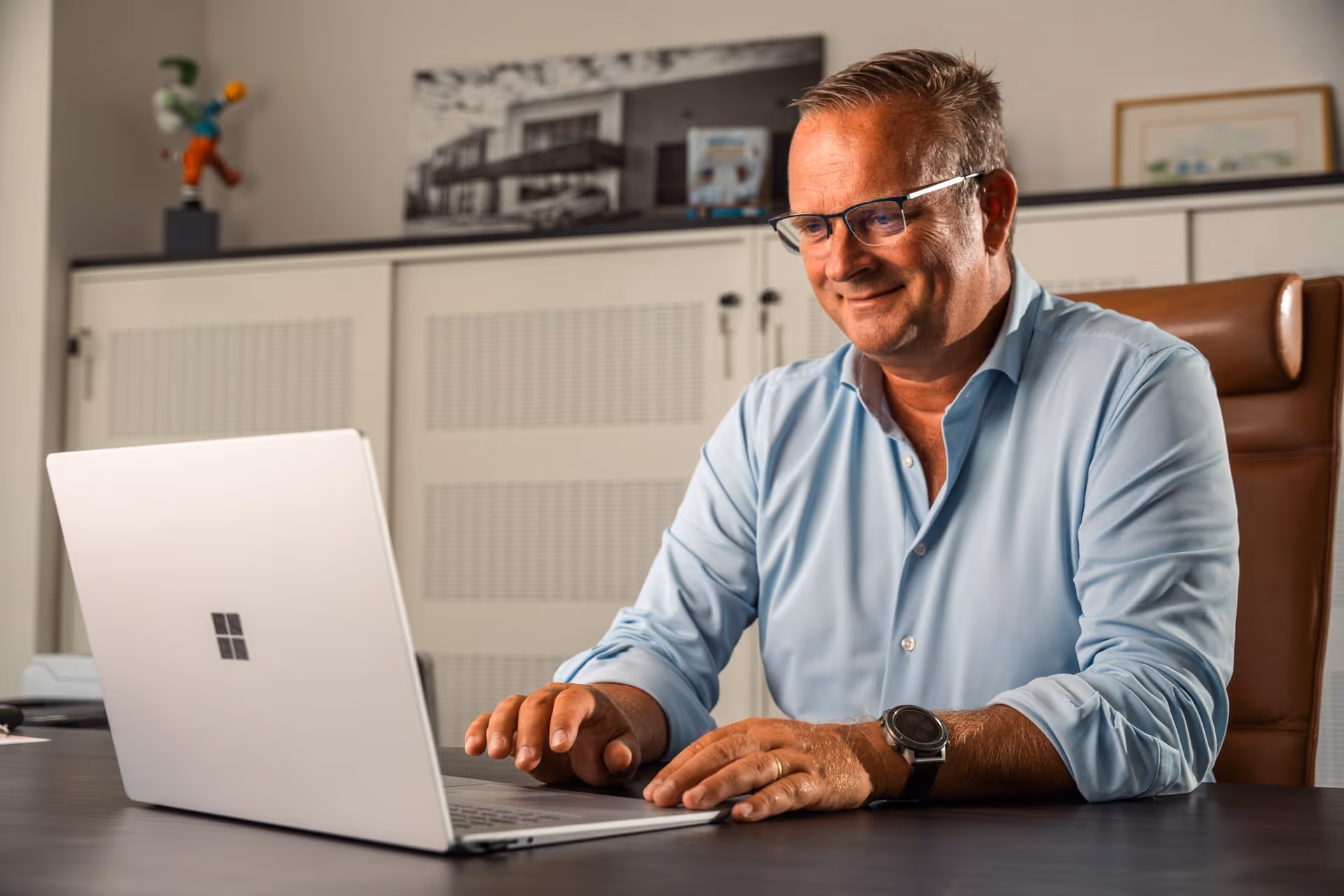 Een glimlachende man met een bril en een blauw overhemd die achter een bureau in een kantooromgeving op een laptop werkt.