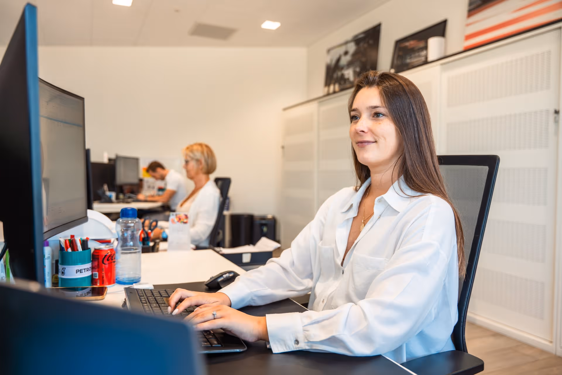 Een glimlachende vrouw in een wit shirt die op een laptop typt in een modern kantoor, met collega’s op de achtergrond.
