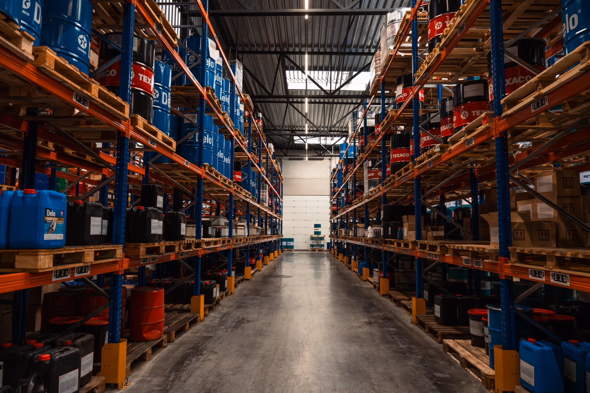 Een gangpad in een magazijn met stellingen vol blauwe en rode olievaten en containers, onder een metalen dak.