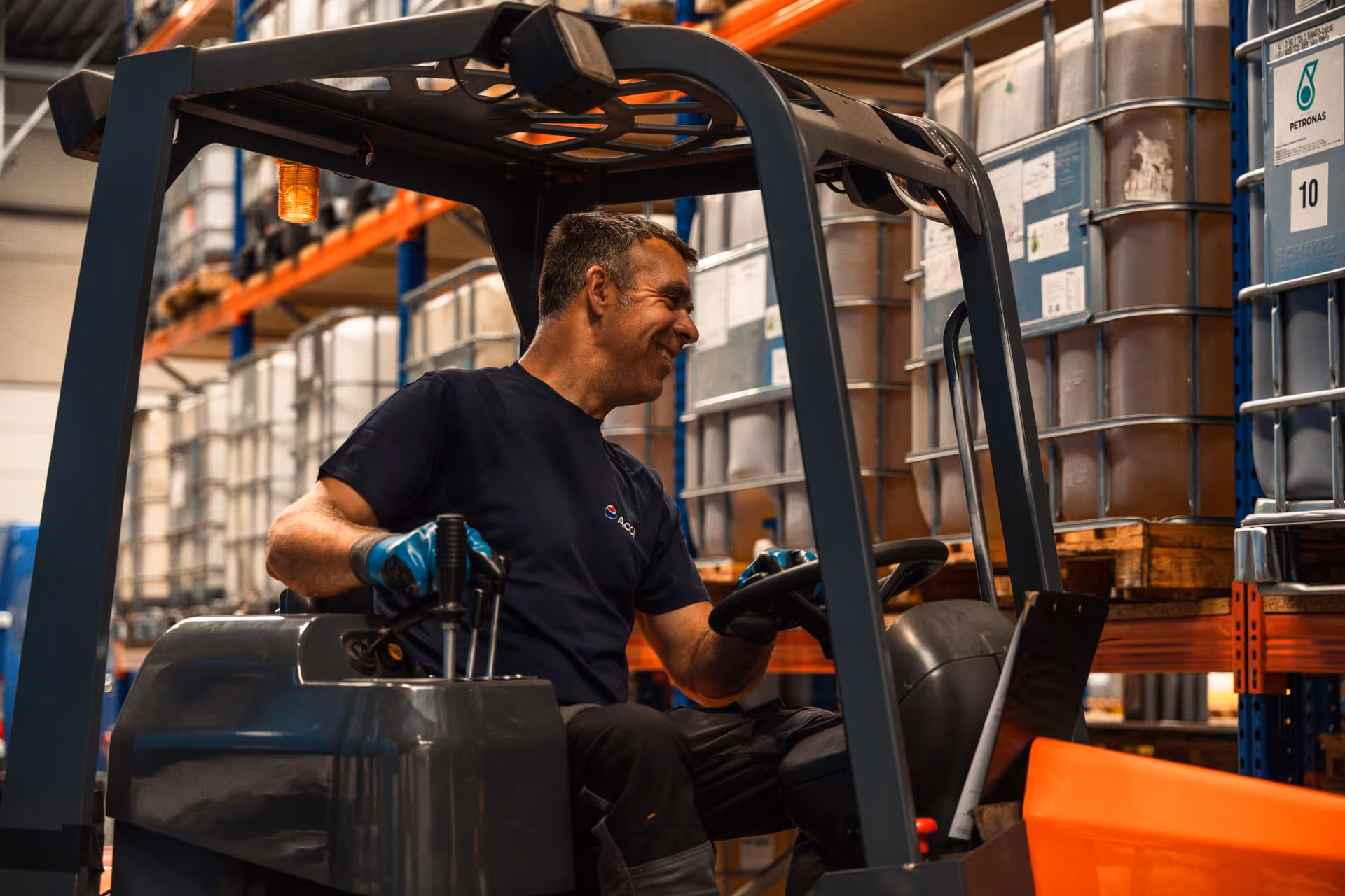 Een magazijnmedewerker die glimlacht terwijl hij met een vorkheftruck tussen de rekken met grote containers rijdt.