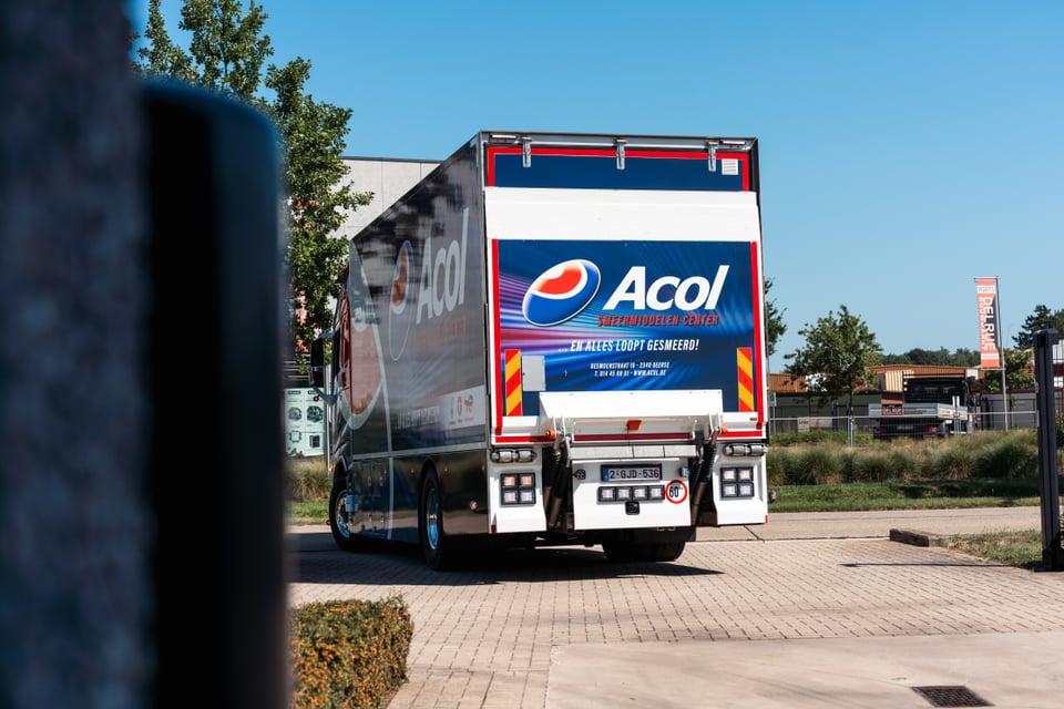 Achterkant van een blauw-witte bestelwagen van het merk Acol, die buiten op een verharde ondergrond geparkeerd staat.