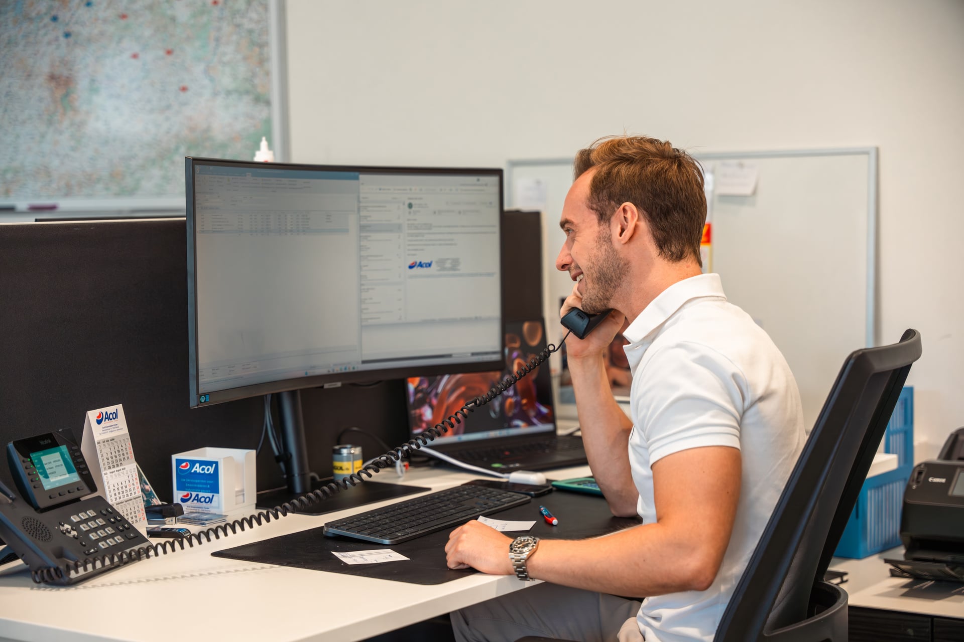 Een man in een wit overhemd zit aan een bureau en is aan het bellen, met computerschermen en kantoorbenodigdheden om hem heen.