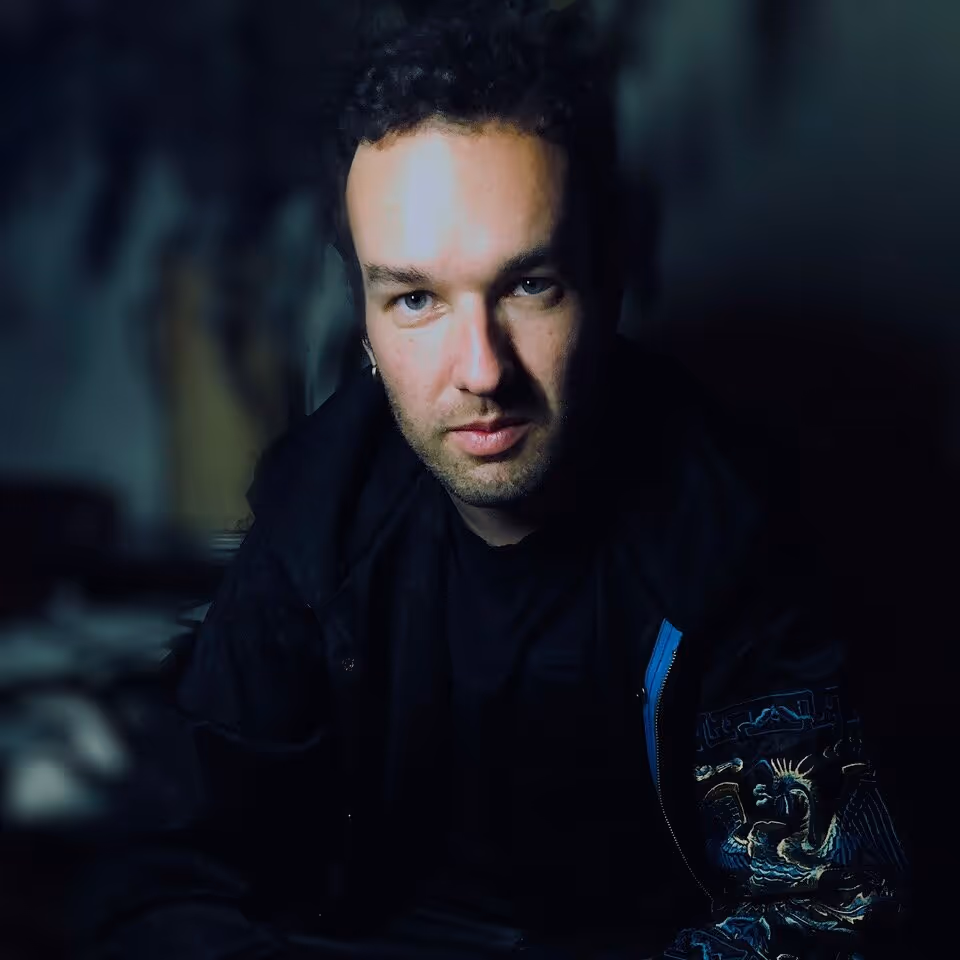 Man with curly dark hair and light skin wearing a black jacket with blue and gold embroidered design on the sleeve, looking directly at the camera in low light.