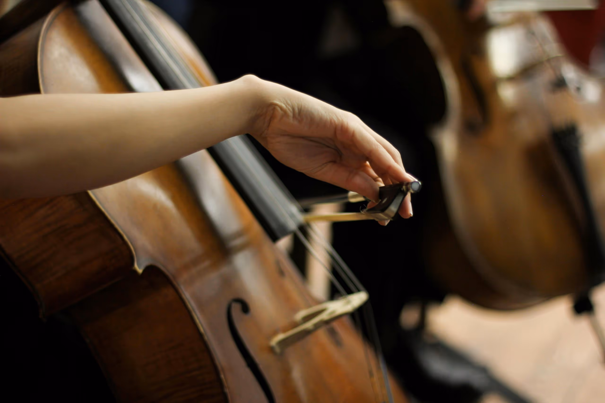 Close-up of a person's hand playing a cello with another cello blurred in the background.