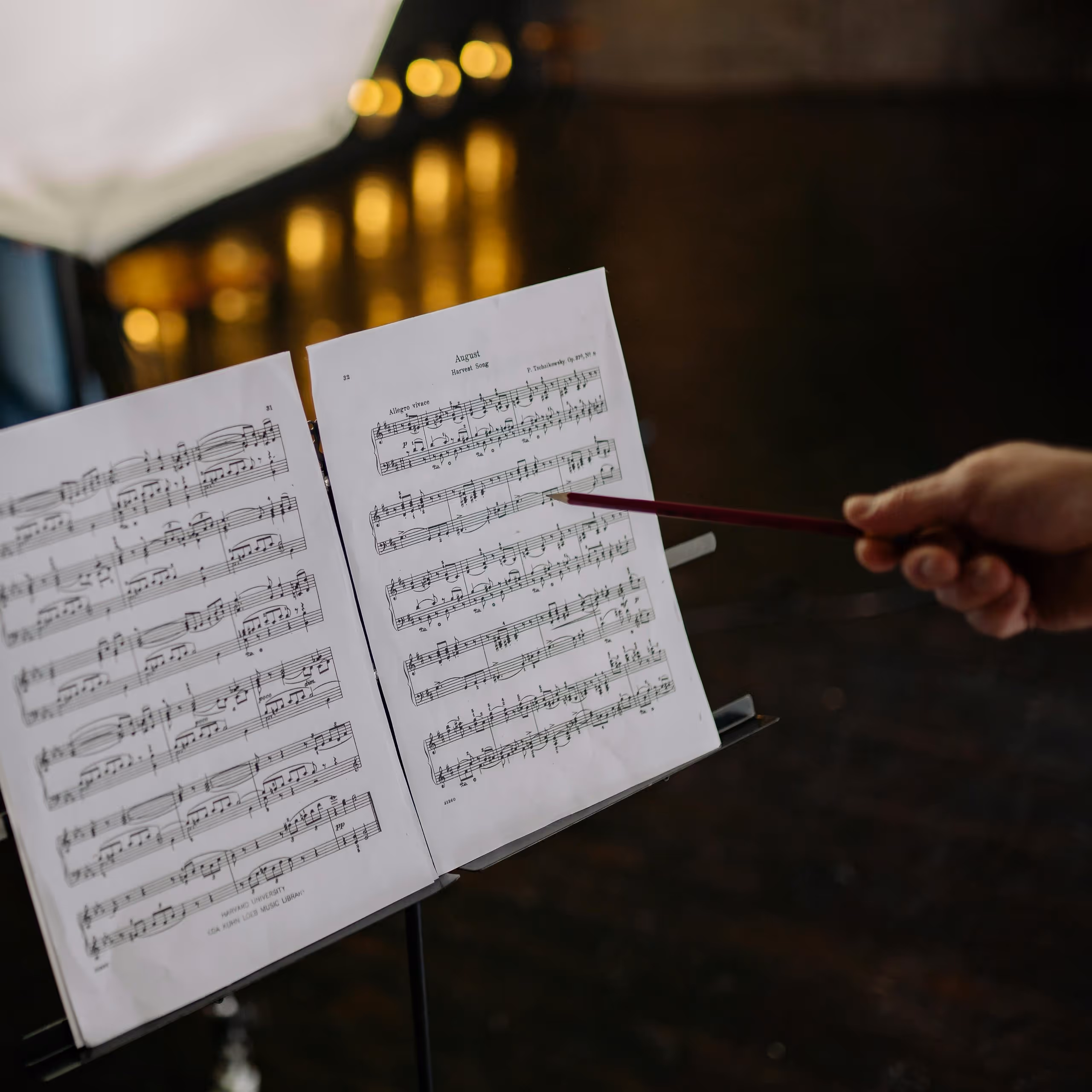 Hand holding a pencil pointing to sheet music titled 'August Harvest Song' on a music stand.