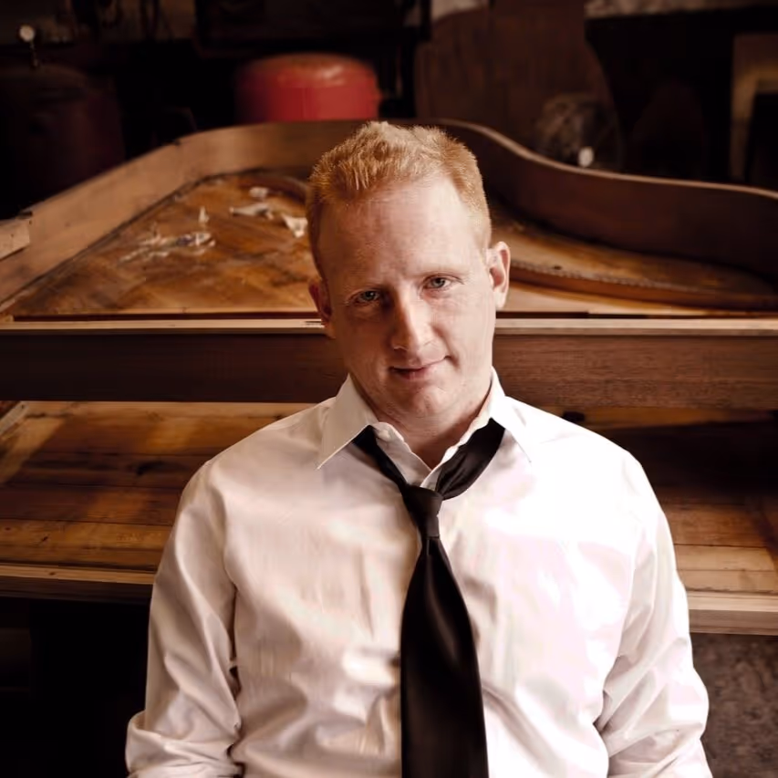 Man with short red hair wearing a white shirt and black tie sitting in front of an old wooden piano frame.