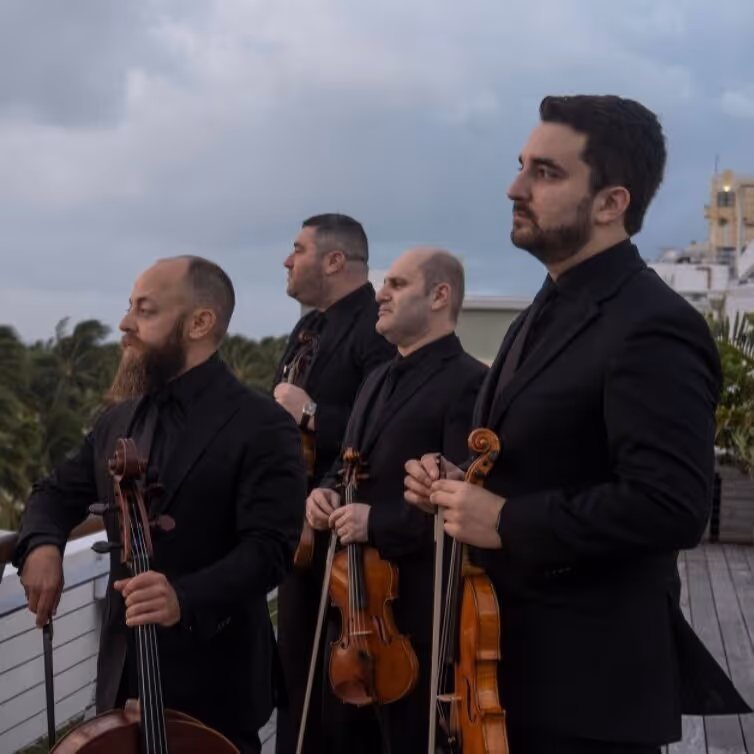 Four men dressed in black suits holding string instruments, standing outdoors on a wooden deck with a cloudy sky and trees in the background.