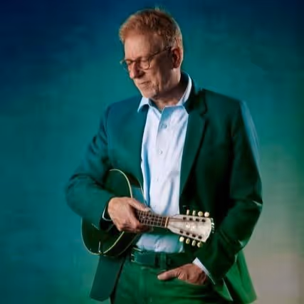 Middle-aged man with glasses wearing a dark green suit playing a mandolin against a gradient green-blue background.