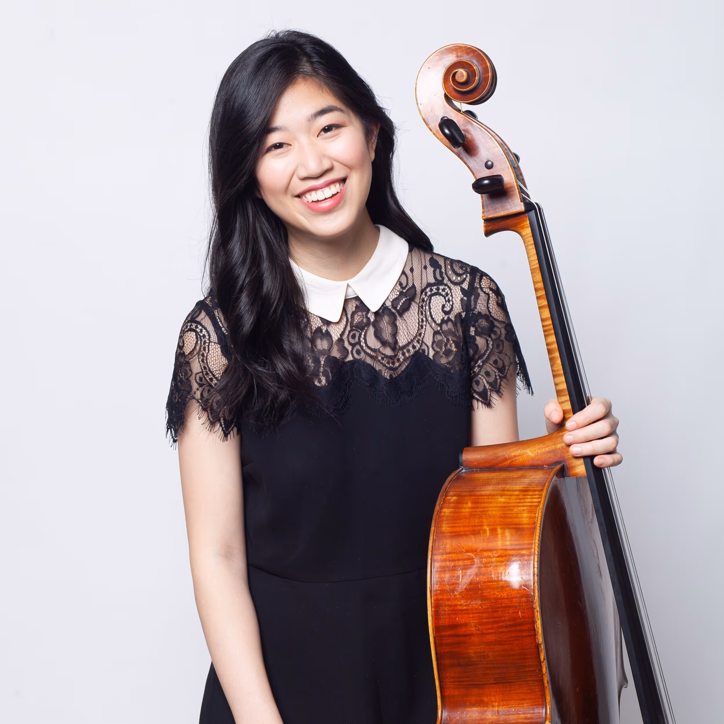 Smiling woman with long black hair in a black dress holding a cello against a white background.