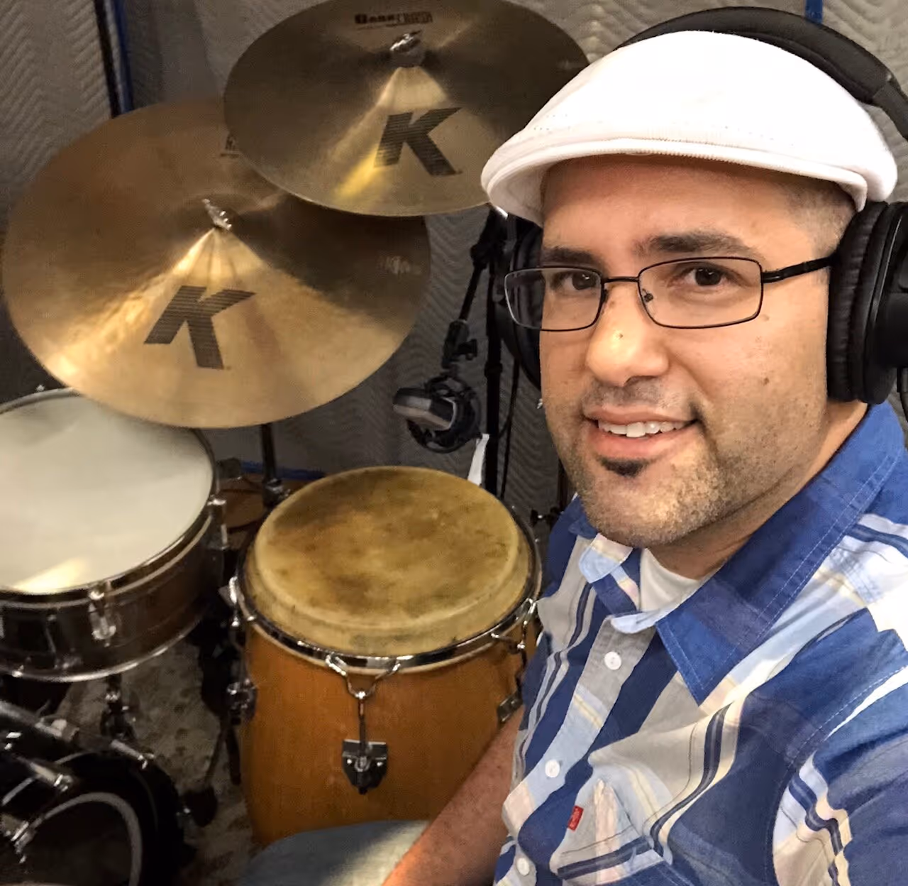 Man wearing a white cap, glasses, and headphones sitting next to a drum set with cymbals and a conga drum.