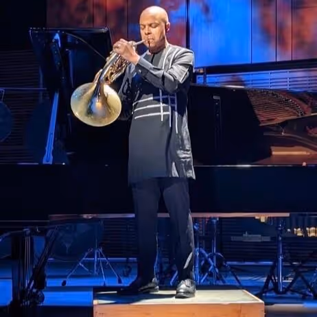 Smiling woman in a black dress with lace details holding a cello against a plain white background.
