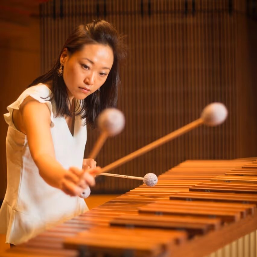 Four men in black suits standing outdoors holding string instruments, including violins and a cello, against a cloudy sky.