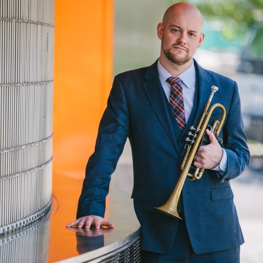 Smiling woman in a black dress with lace details holding a cello against a plain white background.