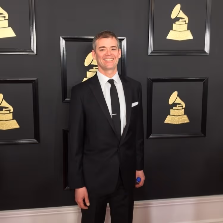 Man with short red hair wearing a white shirt and black tie, seated in front of an old wooden piano with exposed strings.