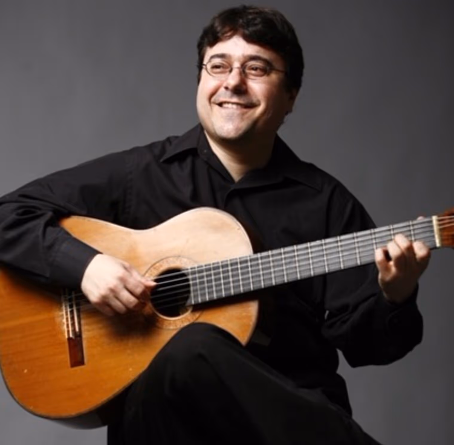 Four men in black suits standing outdoors holding string instruments, including violins and a cello, against a cloudy sky.