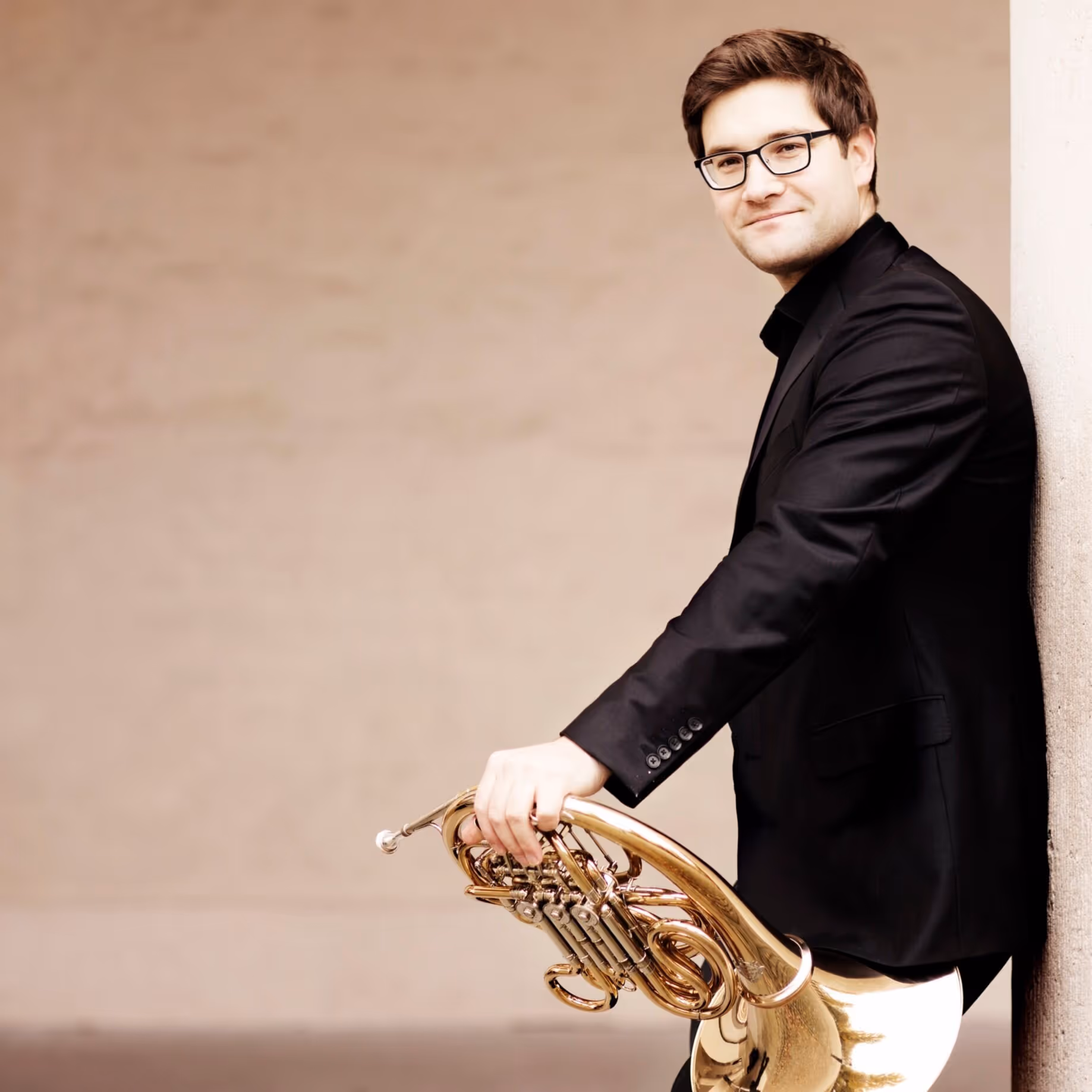 Four men in black suits standing outdoors holding string instruments, including violins and a cello, against a cloudy sky.