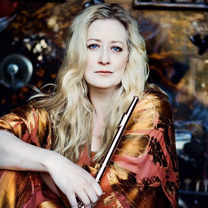 Man with short red hair wearing a white shirt and black tie, seated in front of an old wooden piano with exposed strings.