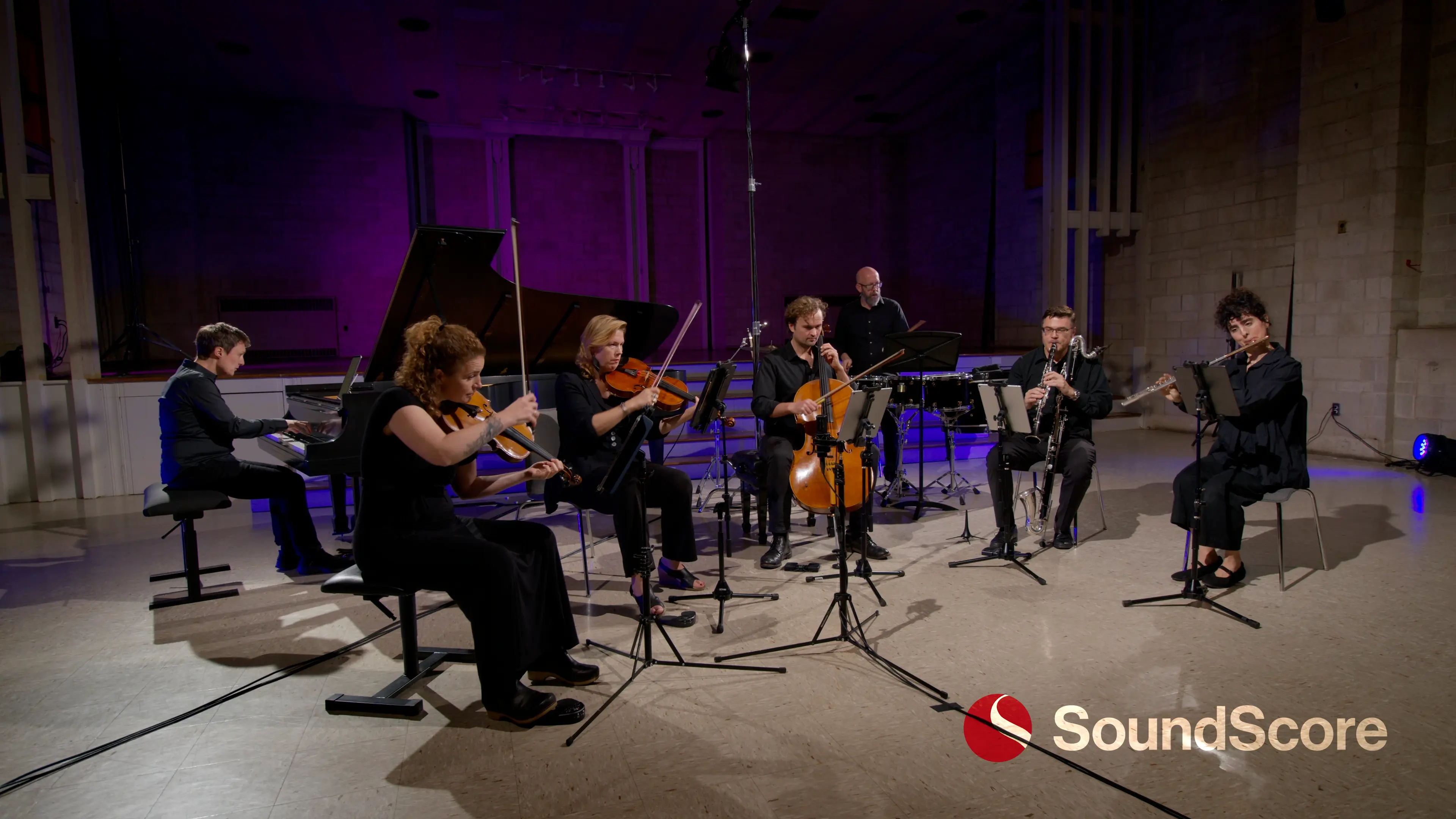 Group of six musicians with string and wind instruments seated and standing in a recording studio with microphones and wood floor.