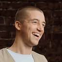 Young man with a shaved head smiling against a dark brick background.