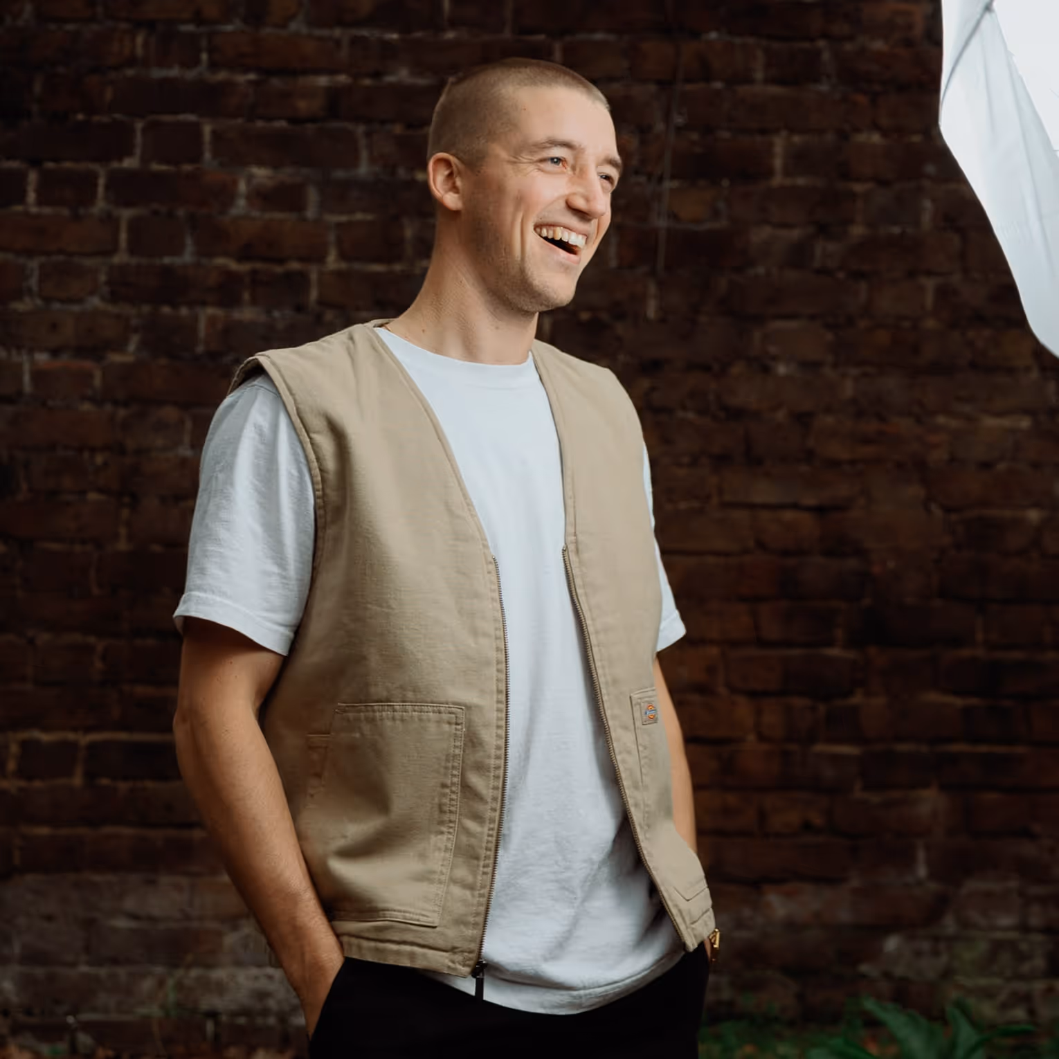 Smiling man with short hair wearing a beige vest over a white t-shirt standing against a brick wall background.
