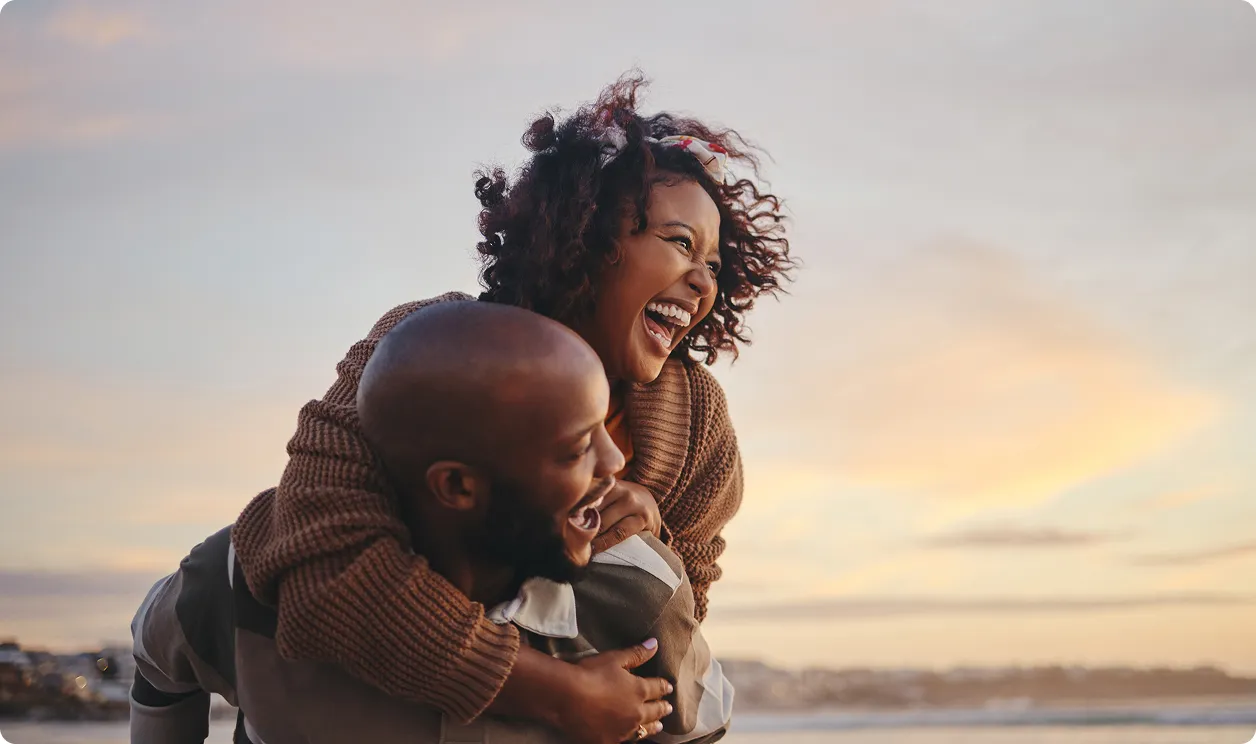 Un homme porte une femme sur son dos alors qu'ils rient tous les deux au bord de la mer au coucher du soleil.