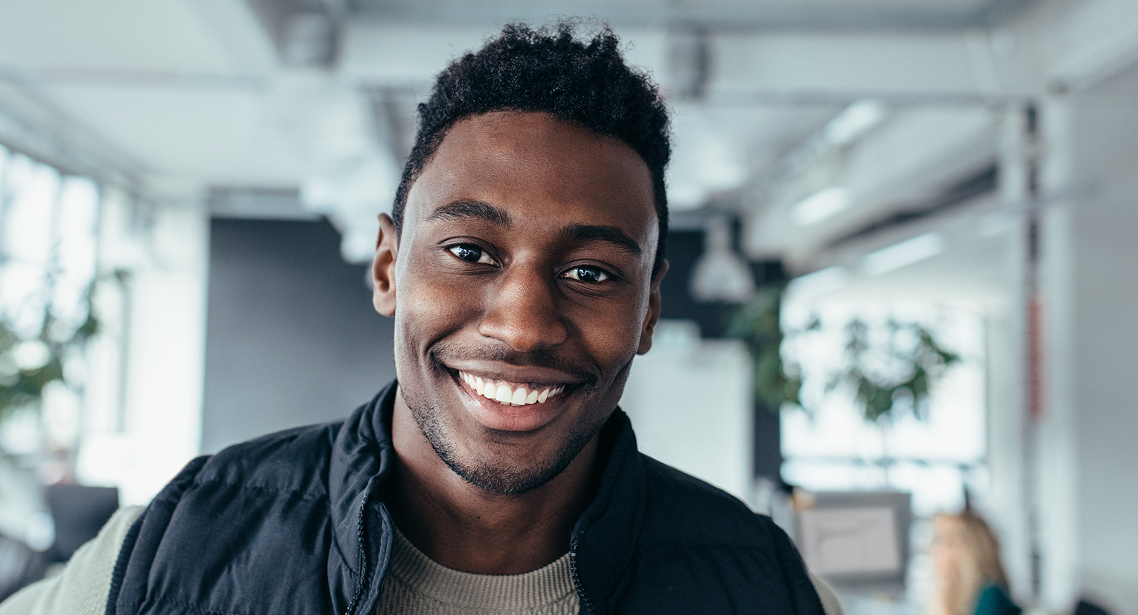 Jeune homme souriant portant un gilet noir dans un bureau lumineux.