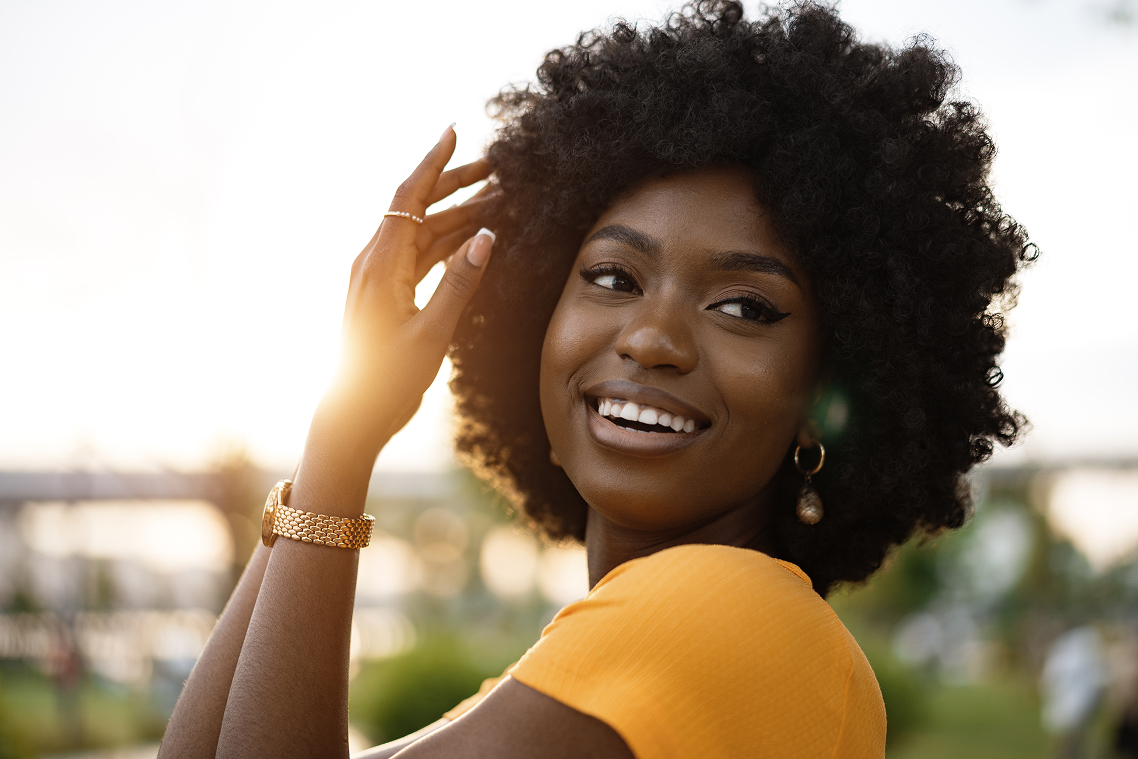 Jeune femme souriante avec cheveux bouclés afro portant un t-shirt orange et des bijoux, touchant ses cheveux au coucher du soleil.