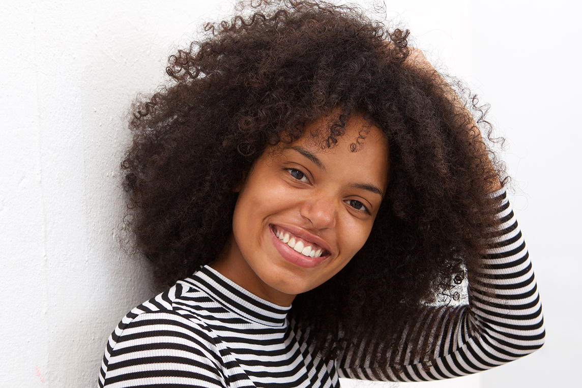 Jeune femme souriante avec de longs cheveux bouclés portant un t-shirt rayé noir et blanc, posant contre un mur blanc.