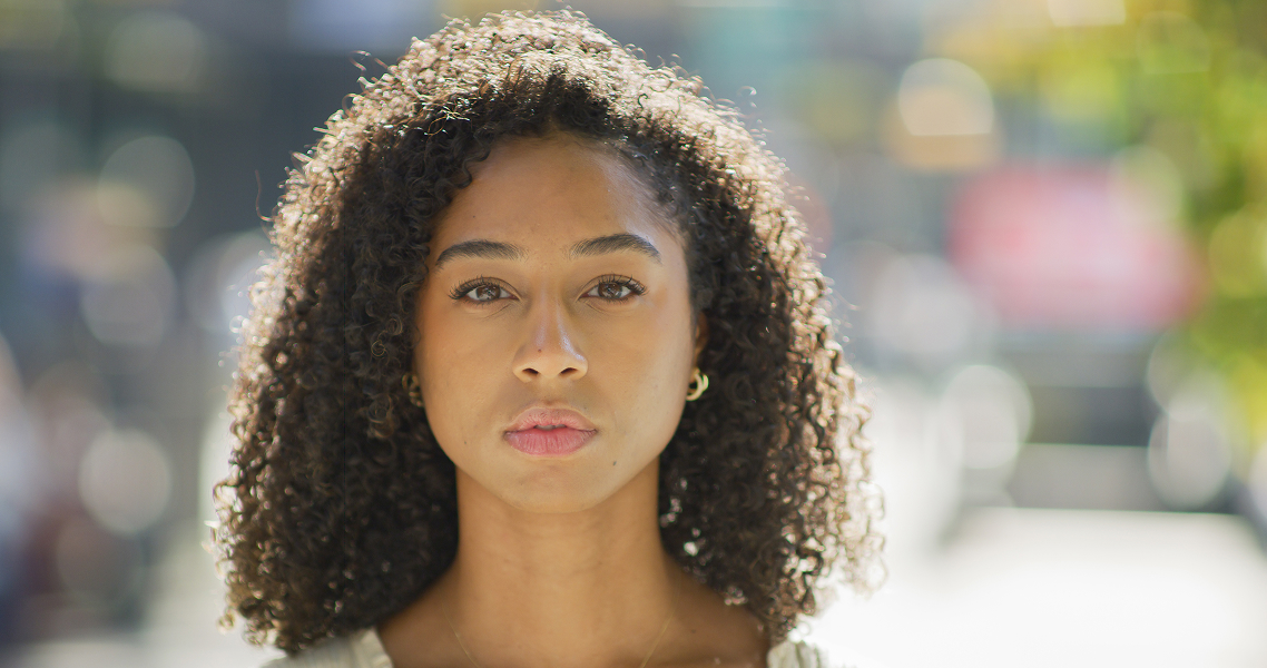 Portrait d'une jeune femme aux cheveux bouclés noirs et expression neutre, en extérieur avec un arrière-plan flou lumineux.