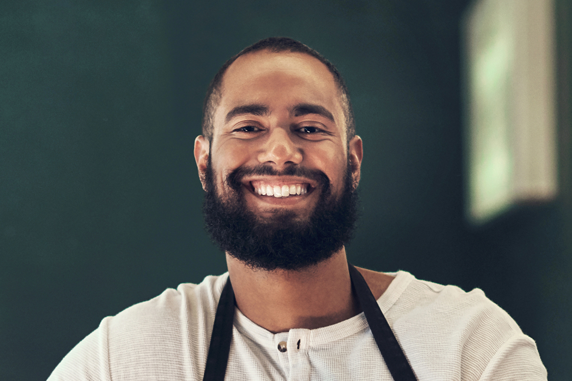 Portrait d'un homme souriant avec une barbe et un tablier noir sur un t-shirt blanc.