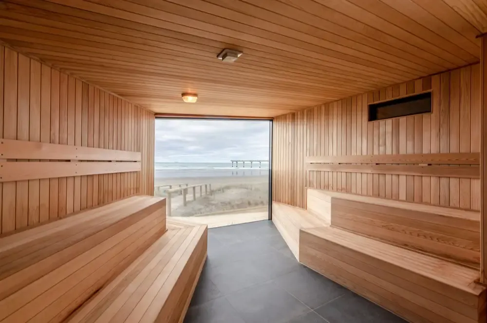 Interior of a wooden sauna room with tiered benches and a large window overlooking a beach and ocean view with a pier.
