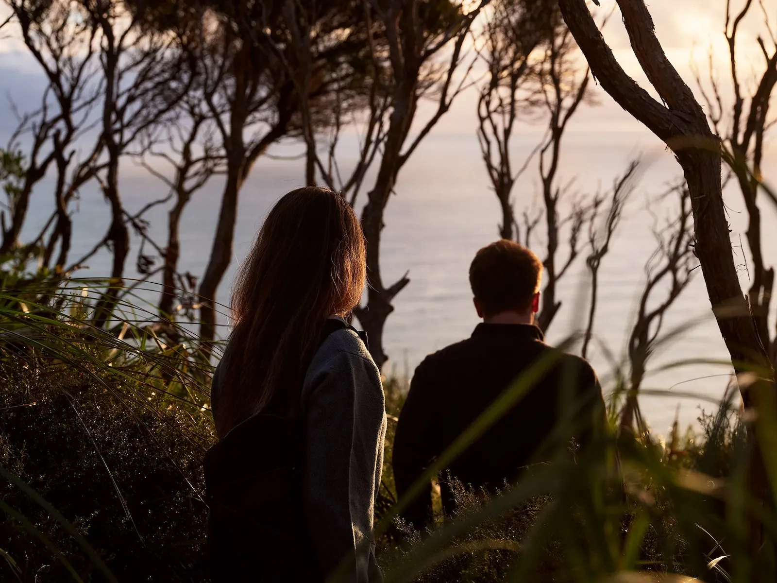 Two people standing among trees looking at the ocean during sunset.