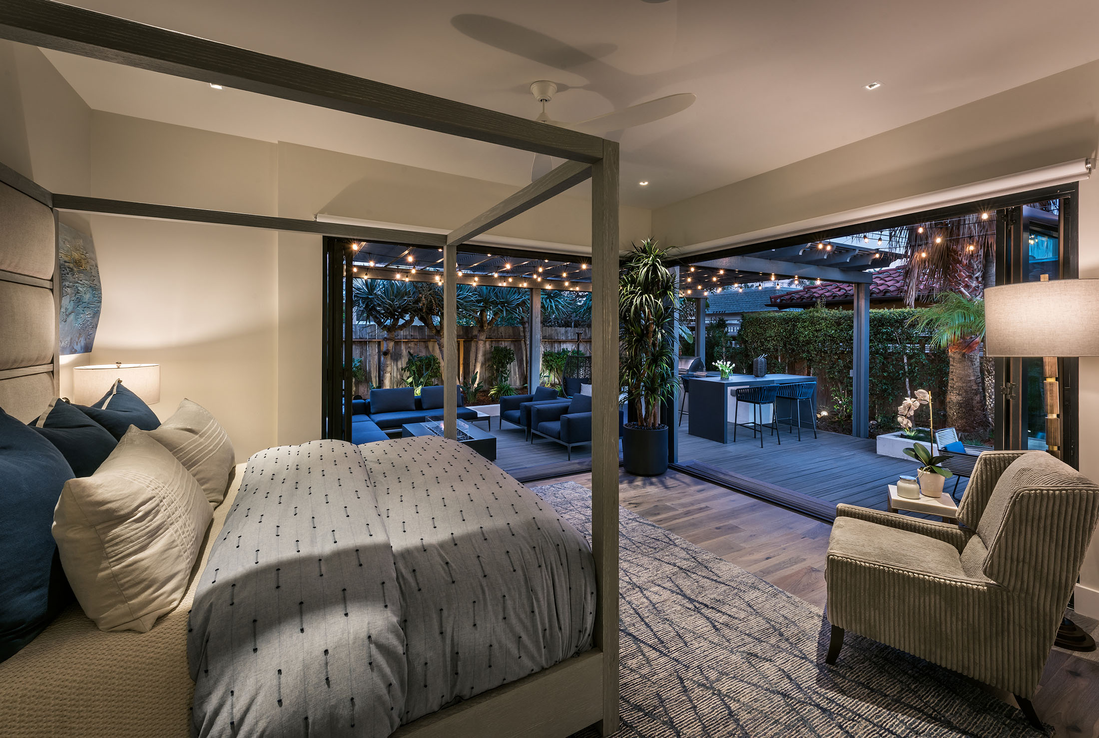 Modern bedroom with a four-poster bed, an armchair, and large sliding glass doors opening to a lit outdoor patio with seating and a dining area.