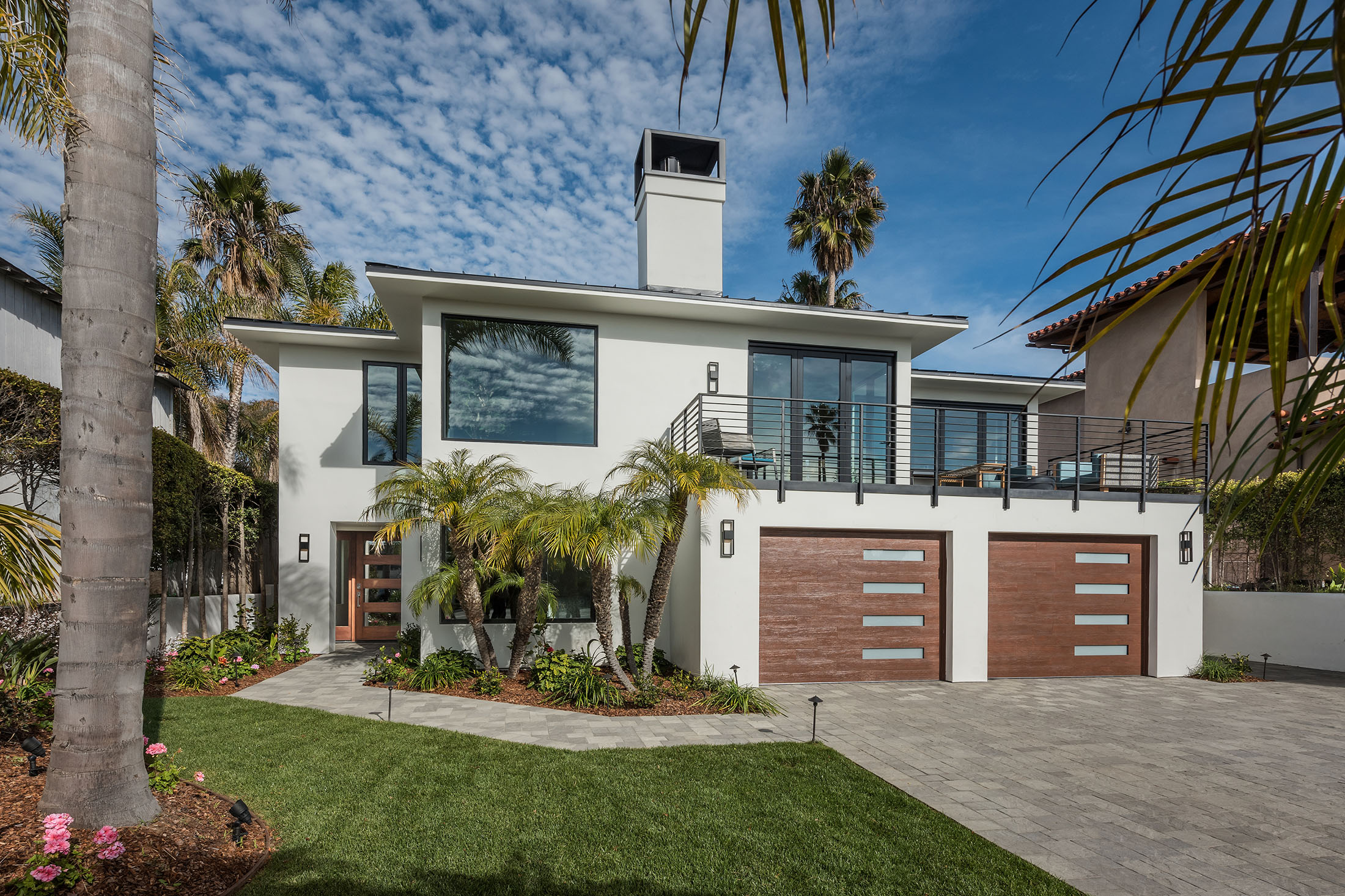 Modern two-story white house with large windows, double wooden garage doors, a balcony with metal railing, and landscaped front yard with palm trees and green lawn.