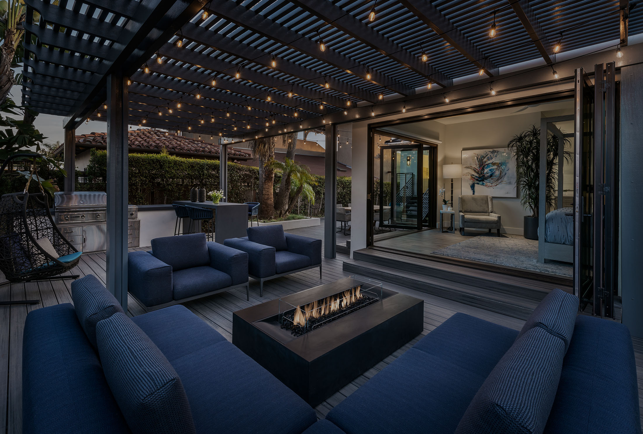 Modern outdoor patio with blue sofas around a rectangular fire pit under string lights, adjacent to an open indoor living space.