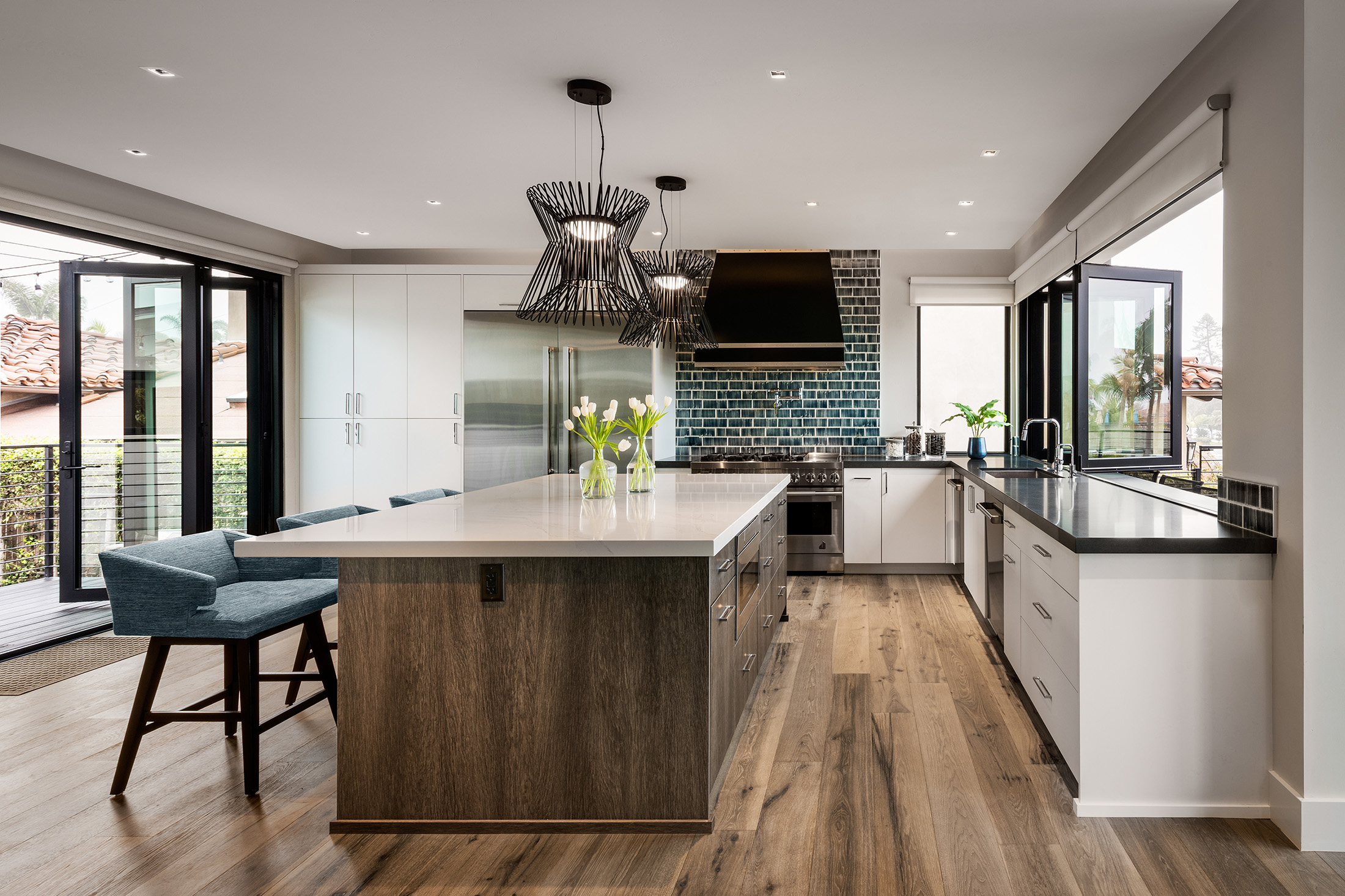 Modern kitchen with large island, blue chairs, pendant lights, stainless steel appliances, blue tile backsplash, and wood flooring.