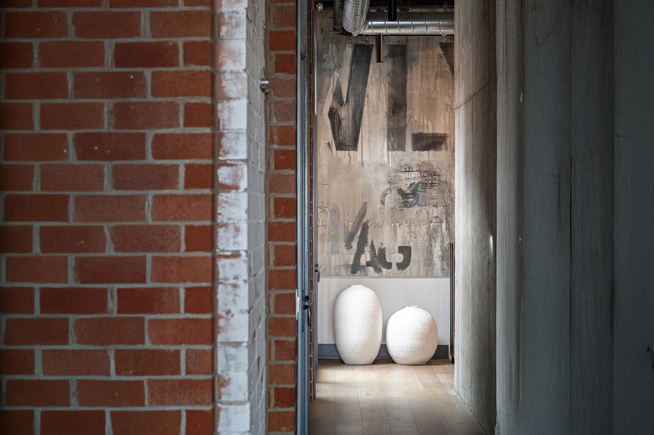 Narrow hallway with exposed brick wall on the left, concrete wall on the right, and two large white vases on a wooden floor against an abstract painted wall.
