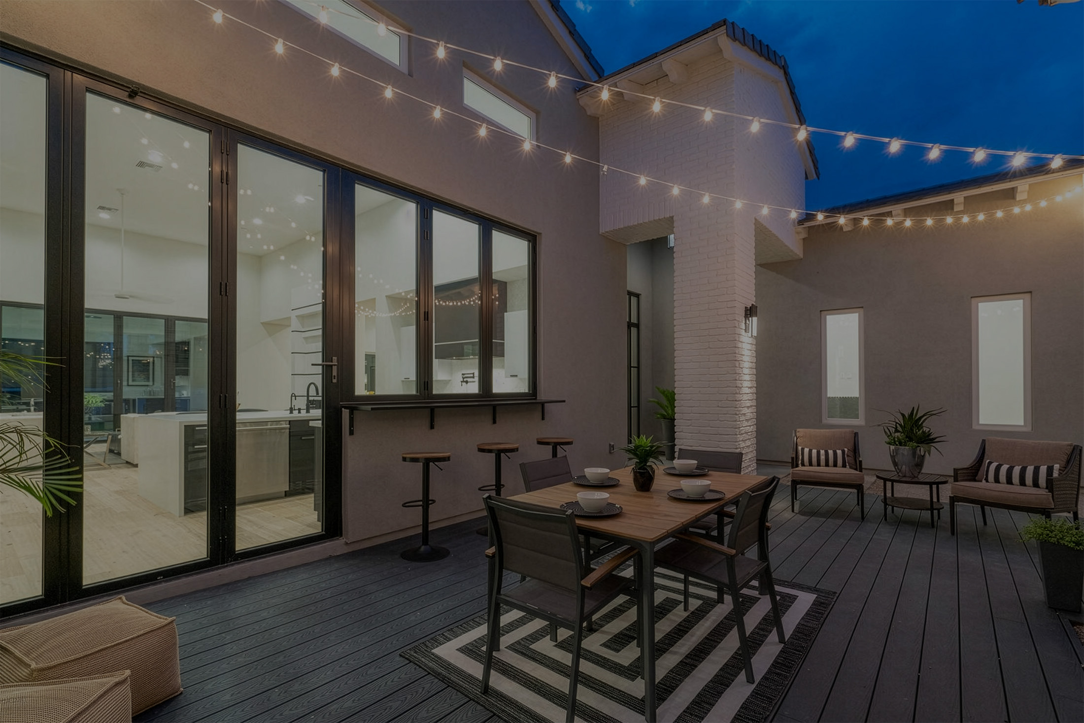 Modern outdoor patio at dusk with string lights, wooden dining table set for four, bar stools, and seating area with chairs and plants.